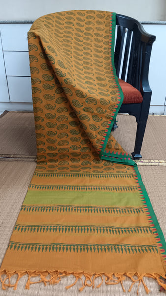 Striking mustard cotton saree featuring a captivating temple border with red and green motifs. Paisley prints in dark green adorn the body, creating a confident and traditionally inspired look.